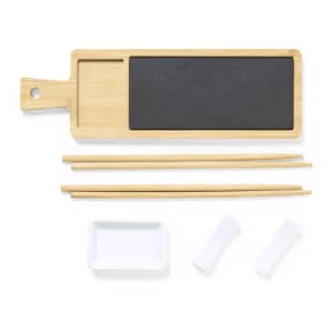 A bamboo sushi serving board with a black stone section, chopsticks, and small white dishes.