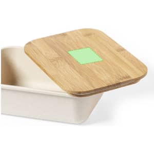 A rectangular bamboo lid sits atop a light-coloured container. The lid features a small green detail and a natural finish.