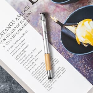 A silver and bamboo stylus touch ball pen resting on a printed menu. The background features a dessert dish.
