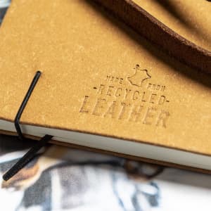 A notepad featuring a tan cover made from recycled leather, with a black elastic closure and a branding logo.