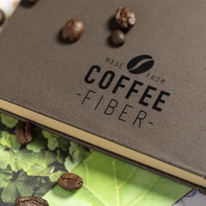 A notepad made from coffee fibre, with a textured brown cover scattered with coffee beans and green leaves.