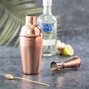 A copper cocktail shaker with a jigger and a gold spoon, set against a grey surface and a bottle of vodka in the background.