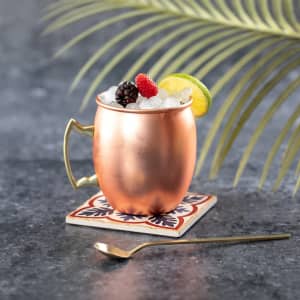 A copper mug with a handle holds a cocktail topped with berries and a lime slice, placed on a decorative coaster.