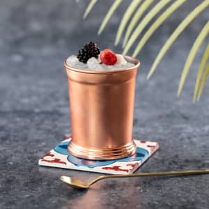 A copper drink cup filled with crushed ice and topped with berries, sitting on a colourful coaster with a gold spoon beside it.