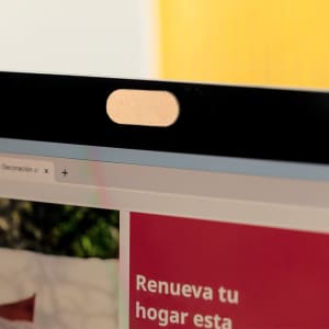 A webcam cover in a rose gold colour, positioned on a laptop screen, with a blurred webpage in the background.