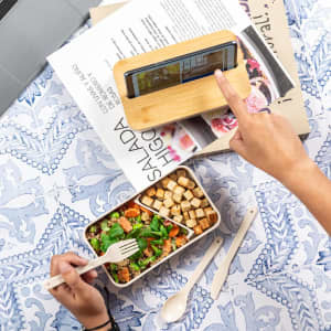 A lunch box with a salad and croutons, beside a phone holder and recipe magazines on a patterned tablecloth.