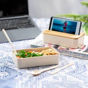 A beige lunch box with a wooden lid, containing salad and snacks, alongside a smartphone stand.