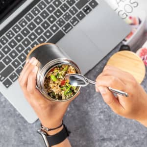 A hand holding a metal lunch container with a mix of quinoa and vegetables, next to a laptop. The container has a wooden lid.