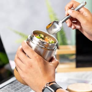 A stainless steel food jar with a wooden base, being filled with colourful food using a spoon.