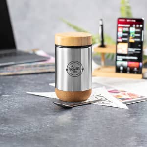 A stainless steel lunch box with a cork base and a wooden lid, featuring a logo. Set on a desk with a laptop and papers.