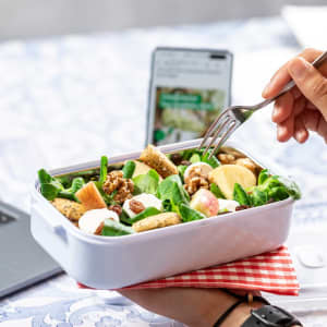 A hand holds a fork over a white thermal lunch box filled with salad, walnuts, and apple pieces, resting on a red gingham cloth.