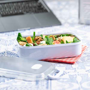 A white thermal lunch box filled with salad, sitting on a red checkered cloth next to a laptop.