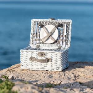 A white wicker picnic basket with a hinged lid, set against a scenic water background. It features an internal compartment and a logo.