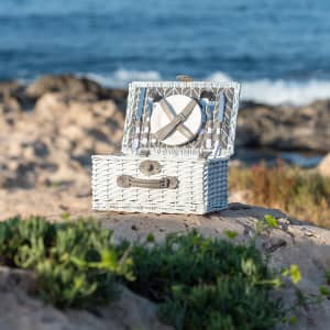A white wicker picnic basket with an open lid, displaying a round plate inside, set against a coastal background.