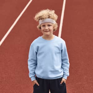 A child wearing a light blue sweatshirt and dark shorts stands on a running track, with a headband in their hair.