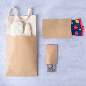 A beige paper bag holds oven mitts, with smaller brown and colourful patterned bags beside it. All items are displayed on a grey surface.