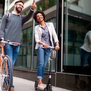 A woman rides a scooter while a man with a bicycle points, both smiling in an urban setting.