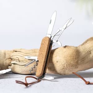 A multifunction pocket knife with stainless steel tools and a cork handle, displayed on a piece of driftwood.