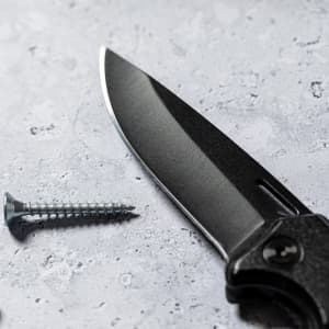 A black Datrak Pocket Knife blade lies beside a silver screw on a textured surface.