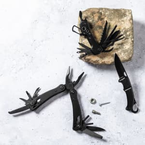 A black multi-tool with various implements rests on a stone, alongside a black pocket knife and metal fasteners.