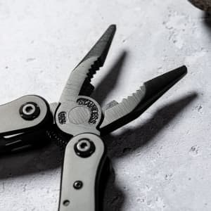 A multitool with black metal finishing, featuring pointed and serrated pliers, placed on a textured surface.