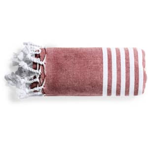 A folded towel pareo in a pinkish-red hue with white stripes and tasselled edges. It features a branding logo.