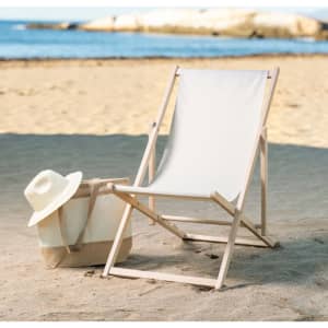 A beige leisure chair with a wooden frame beside a canvas bag and straw hat on a sandy beach with water in the background.