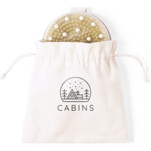 A round exfoliating massage brush in a cotton drawstring bag, featuring a textured surface and a logo.