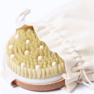 An exfoliating massage brush with soft bristles, featuring a wooden handle and a cream cloth bag for storage.
