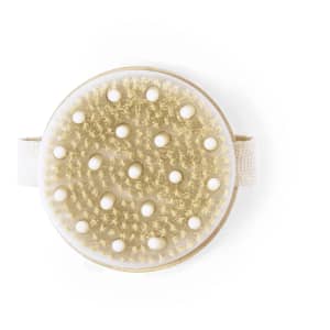 A circular exfoliating massage brush in cream and gold, featuring a textured surface and an elastic strap.