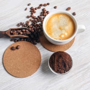 A coffee cup on cork coasters, surrounded by coffee beans and ground coffee in a clear dish.