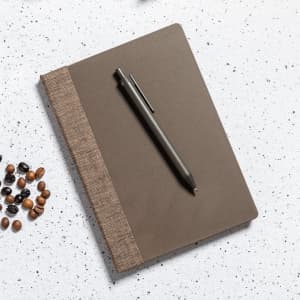 A brown notebook with a textured cover and a grey Bropex pen placed on a speckled surface with coffee beans.