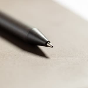 A black pen with a fine tip rests on a neutral-toned surface, featuring a simple design and a branding logo.