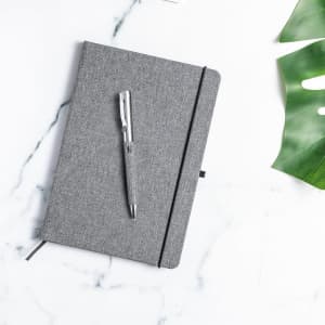A grey notebook with an elastic band and a silver metal pen placed on a marble surface alongside green foliage.