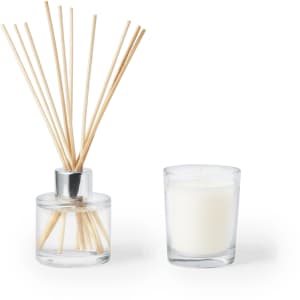 A glass diffuser with reeds alongside a small glass candle, both featuring a white colour. The diffuser has a metallic top.