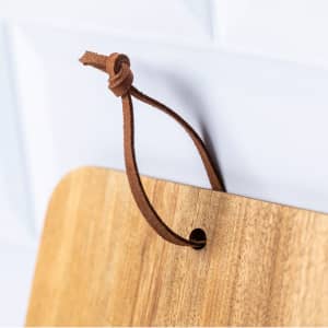 A wooden cutting board with smooth edges, featuring a leather hanging strap attached to a hole at the top.
