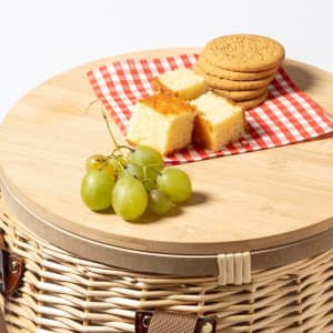 A wicker picnic basket with a wooden lid, featuring green grapes, cake pieces, and biscuits on a checkered cloth.