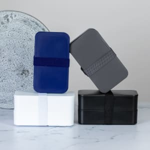 A collection of four lunch boxes in navy, grey, white, and black, stacked creatively against a textured background.