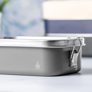 A grey stainless steel lunch box with a metal latch, set against a blurred background.