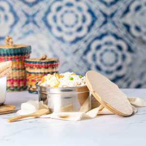 A stainless steel lunch box with a wooden lid, containing rice and vegetables, set on a textured background.