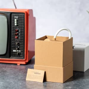 A boxed lamp in white and brown packaging beside an orange retro television on a grey surface, featuring decorative elements.