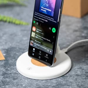 A wireless charger with a white base and cork detailing, holding a smartphone displaying a music app.