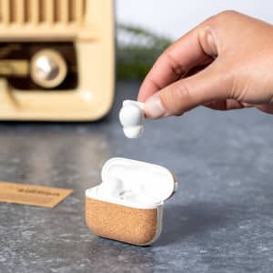A pair of white earphones being held above a cork case, set against a grey surface and a vintage appliance in the background.