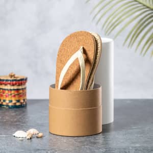 A pair of cork flip flops with cream straps nested in a cylindrical cardboard container.