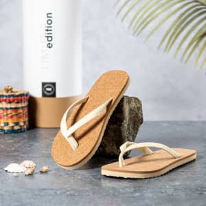 A pair of beige Yassir flip flops with cork soles, positioned on a rocky surface beside shells and a drink bottle.