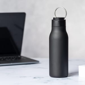 A drink bottle in matte black finish with a lid featuring a metal ring, positioned near a laptop on a marble surface.
