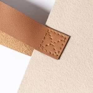 A close-up of a beige tote bag featuring a brown leather strap with decorative stitching. The bag has a textured finish.