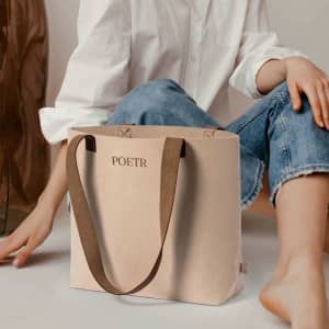 A beige tote bag with brown straps, featuring a logo on the front, placed beside a person in casual attire.