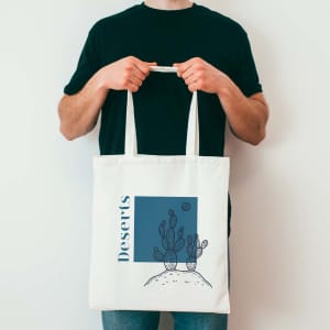 A cream tote bag with cactus illustrations and a blue square design, held by a person in a black shirt.