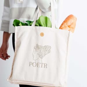 A cream tote bag with a logo, carrying fresh bread and greens, held by a person in a striped top.